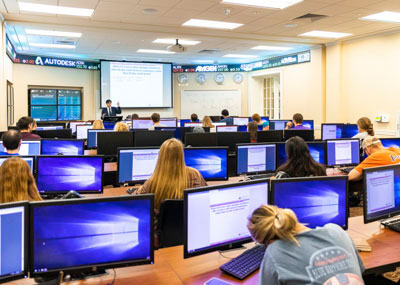 Taken from the back of the trading room classroom with stuednts sitting at computers and a professor presenting with the stock tickers running at the front of the room