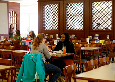 Two female students sitting across the table from one another in a dining hall while having a meal. Other students mingling in the background.
