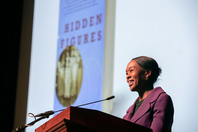 Photo of Margot Lee Shetterly speaking to the crowd with an image of the cover of her book, Hidden Figures, in the background
