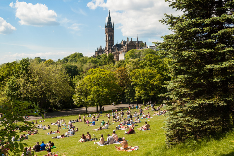 Photo Kelvingrove Park full of people enjoying the Scottish summer with the main building of the University of Glasgow on the top of a hill in the distance.
