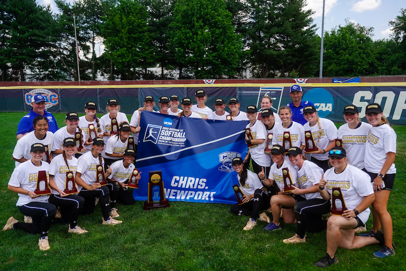 The women's softball team and coaches pose with the championship trophy and banner on the field at Salem, Virginia