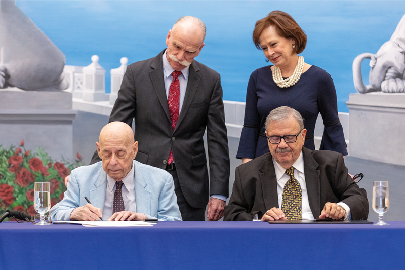 A photo of Dr. Theodore Reiff, seated, wearing a blue jacket, and Dr. Anthony Santory, seated, wearing a brown jacket, signing the agreement to establish the Reiff-Santoro Professorship in History, while Dr. David Doughty, standing, wearing a gray jacket, and Adelia Thompson, standing, wearing a blue dress, look on.