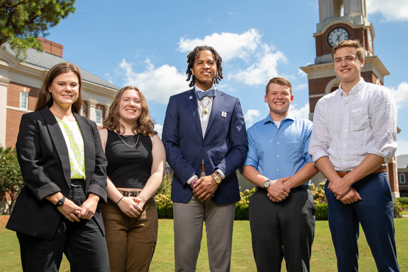 Five individuals stand in front of the campus bell tower on a sunny day.