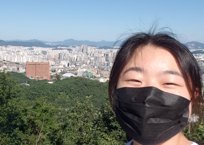 A student wearing a mask stands in the foreground with Seoul, South Korea in the background.