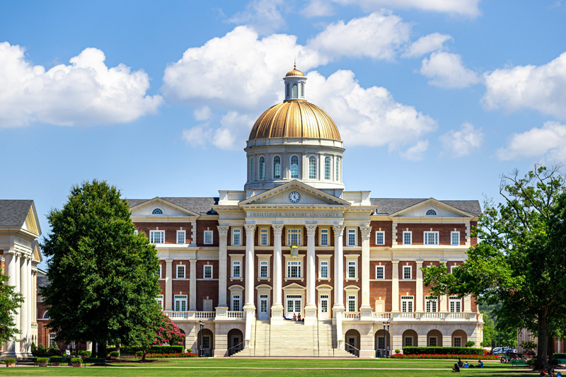 Christopher Newport Hall with a blue sky and clouds behind it and students walking campus and seated on the Great Lawn in front of it.