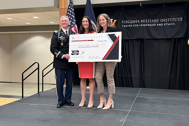 Two students stand beside a man in military uniform. All three are holding a big check with the students' names on it.