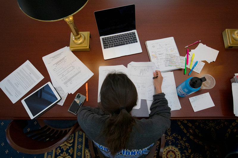 A bird's eye view of a student studying for exams surrounded by papers, their tablet and their laptop.