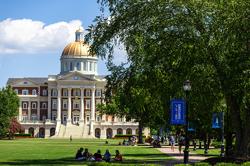 A group of students sit on the Great Lawn near a lamp post with a banner that reads 'Lead A Life of Significance' and in the distance is Christopher Newport Hall.