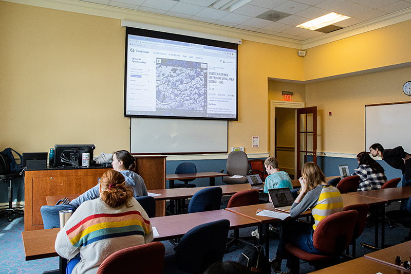 Students sit in a classroom working on laptops while a screen is projected at the front of the room.