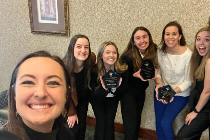 A group of President's Leadership Program students pose with advisor Lacey Grey Hunter while holding three trophies from the Leadership Challenge Event in Kansas.