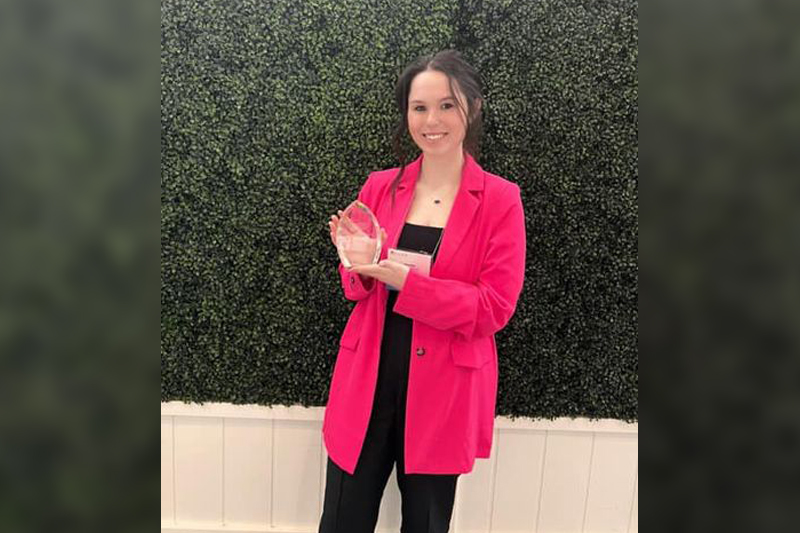 Katherine Paulikonis wears a bright pink blazer while holds her Social Work Student of the Year award in front of shrubbery.