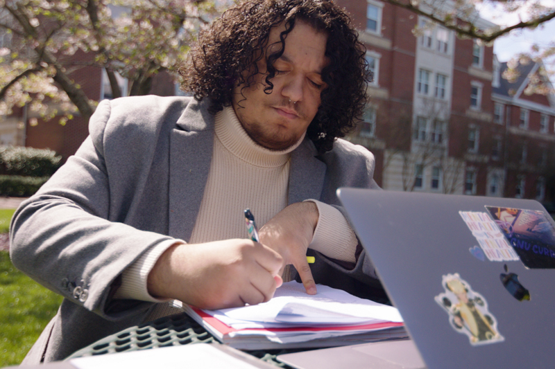 Matthew Johnson '24 sits outside and writes on a paper positioned in front of his laptop.