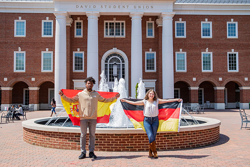 Two students stand in front of a fountain in front of the David Student Union. The male student on the left holds up the flag of Spain behind him and the female on the right holds up the flag of Germany.