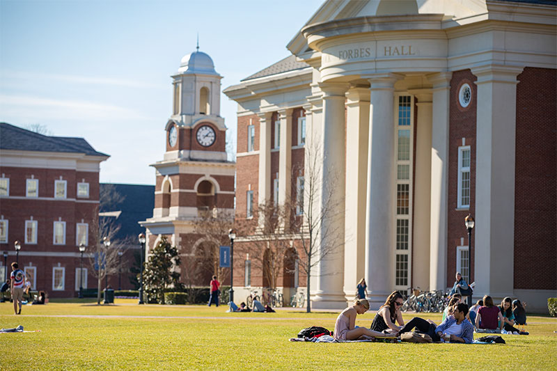 A group of students sit on the Great Lawn with Forbes Hall and a bell tower in the background.