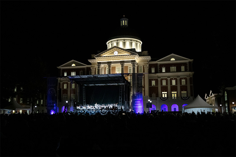 Christopher Newport Hall stands tall at night in the background. In the foreground, the Virginia Symphony Orchestra perform.