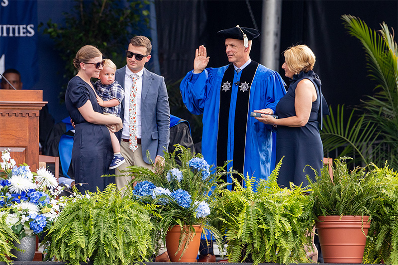 President Kelly raises his right hand and has his left on a Bible held by his wife Angie. Kelly is wearing blue robes with a black stole. The Kellys son Patrick, daughter-in-law Ellen and grandson Liam stand to the left and look on as he takes the oath.