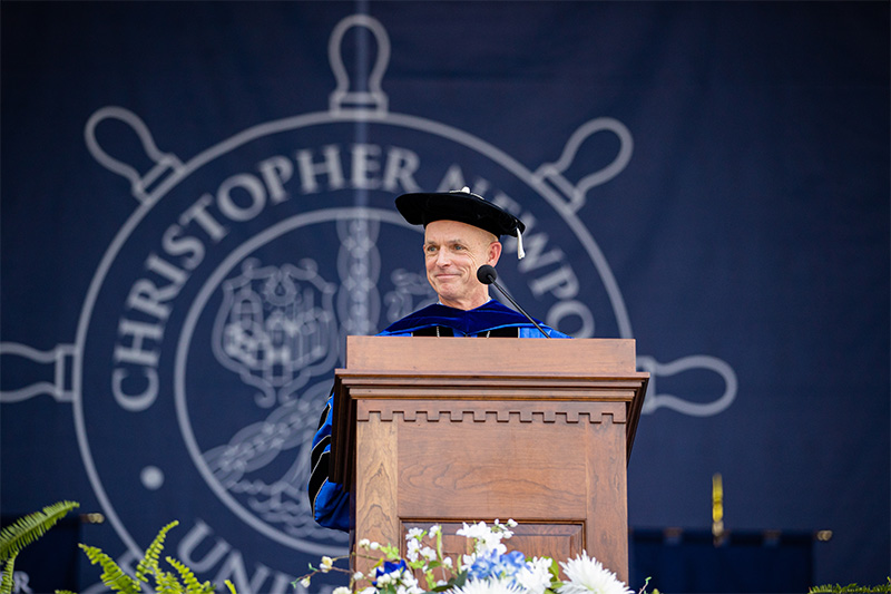 President Kelly stands at a podium wearing a black hat, blue robes and a black stole. A partial view of Christopher Newport's seal can be seen behind him.