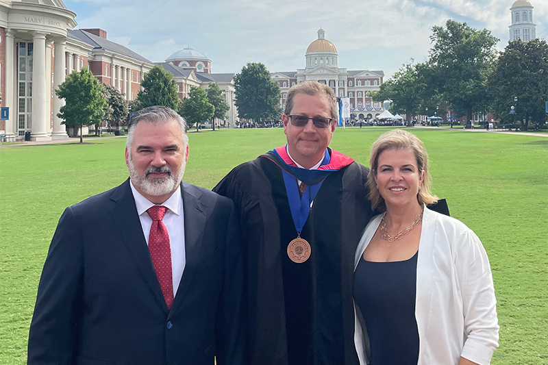 Professor Nathan Busch wears academic graduation regalia whiel standing in between Jim (left) and Cynthia (right) Crawford on the Great Lawn.