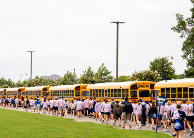 Hundreds of students wearing lilac colored shirts walk towards school buses.