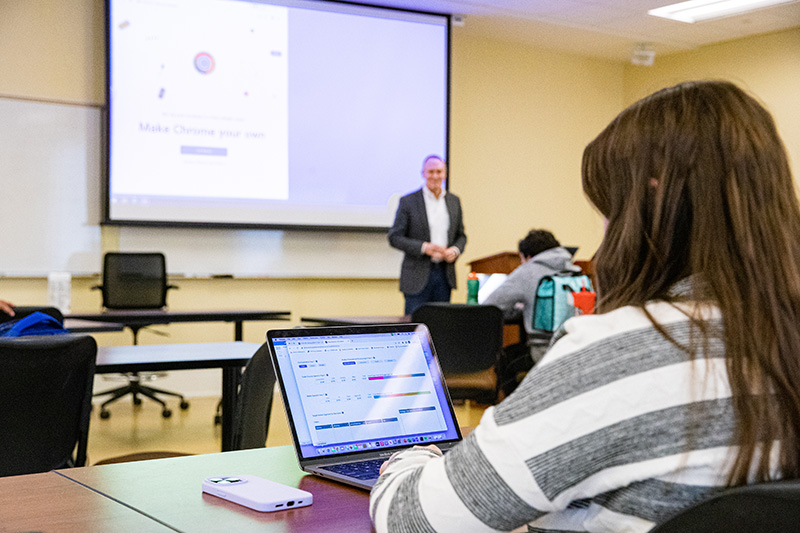 A student's laptop screen is in focus while she pays attention to her professor at the front of the room who is out of focus.