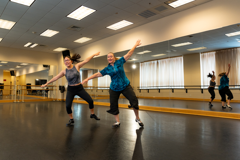 Two individuals tap dance in an empty dance studio.