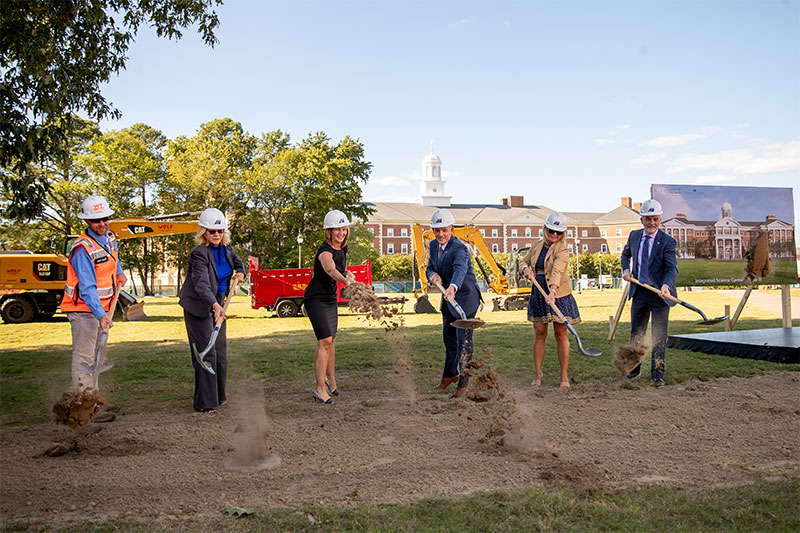 Six individuals are caught mid-action of tossing dirt from their shovel as they break ground.