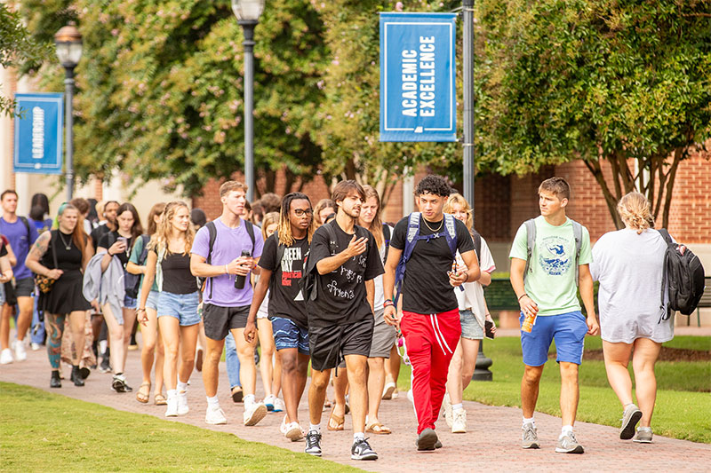 A large crowd of students walk on a sidewalk on Christopher Newport University's campus.