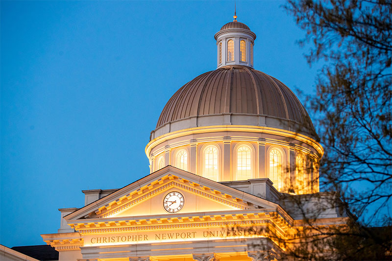 Illuminated cupola of Christopher Newport Hall at dusk. Christopher Newport University is engraved in the stone below the clock.