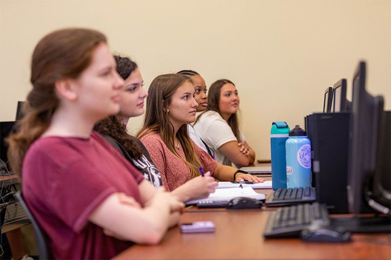 Students sit at a desk with computers in front of them.