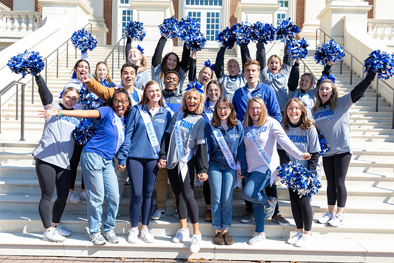 Spirit of CNU finalists wear white sashes and are in expressions of cheer while being surrounded by CNU cheerleaders who have their blue and silver pom poms up to surround the group.