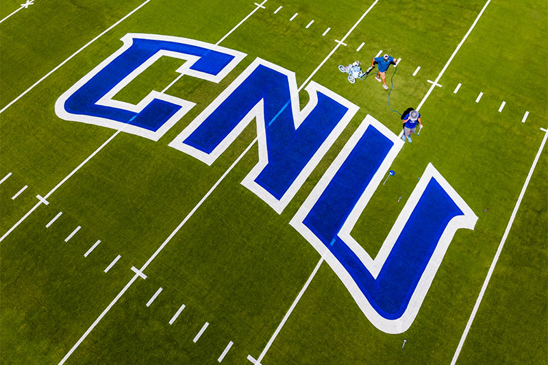 The letters CNU are painted blue and outlined in white by two individuals who stand above the letters. The image is an aerial shot of the football field.
