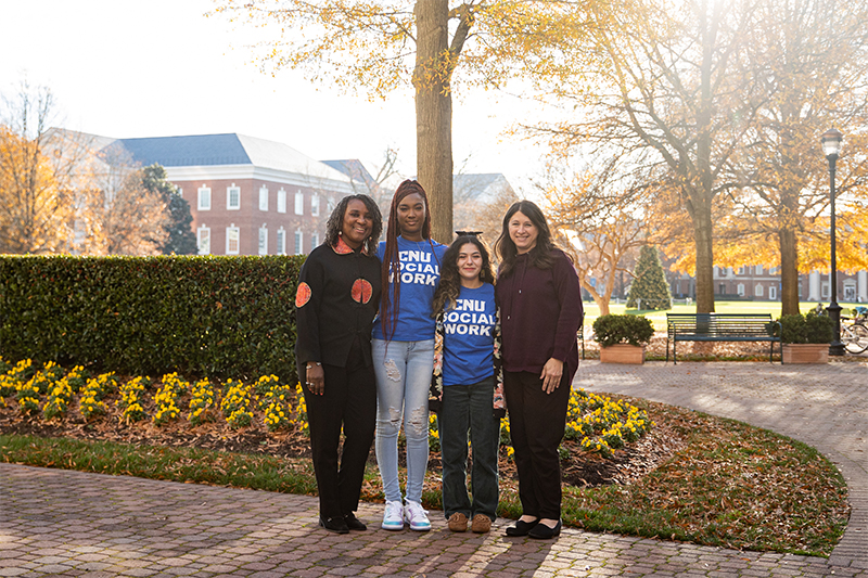 Four women stand together on campus. Two in the middle are wearing CNU Social Work shirts.