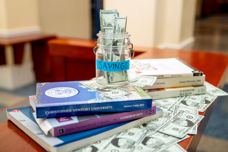 A clear jar labeled Savings sits on top of various books with money underneath.