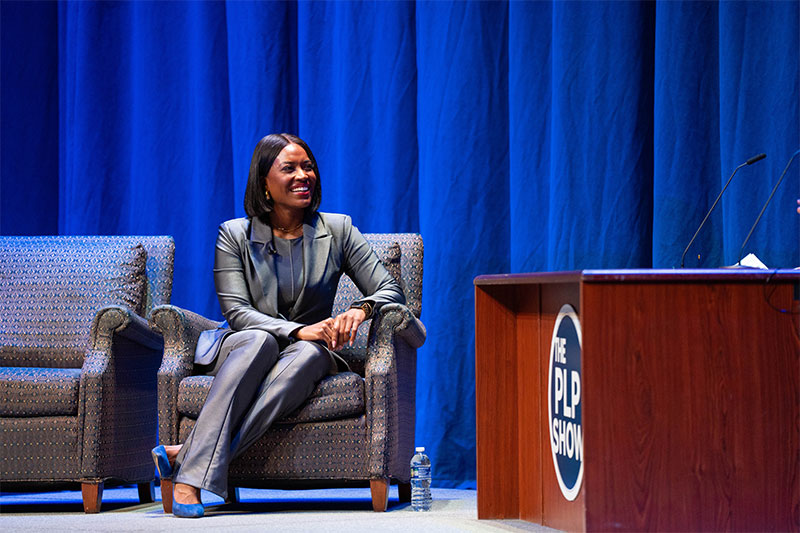 Alencia Johnson sits in a chair in a metallic gray suit on stage. 