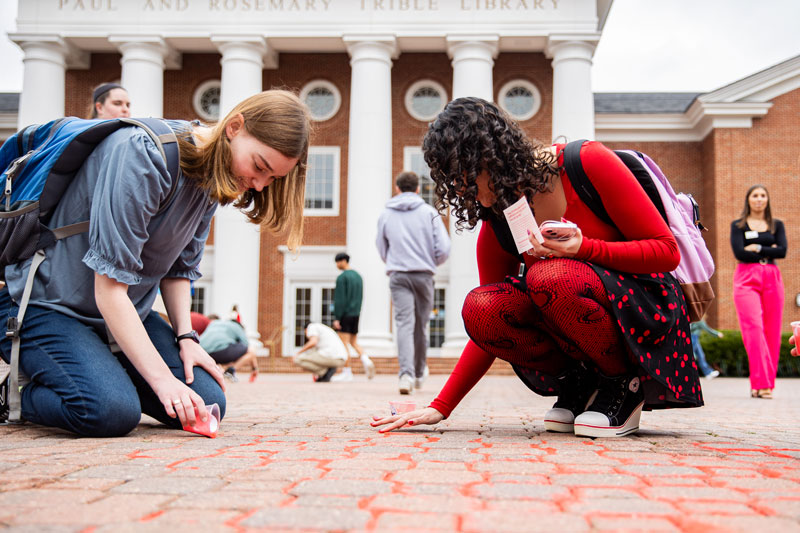 Two students pour red sand from cups into the crevices found between bricks in the Trible Plaza.