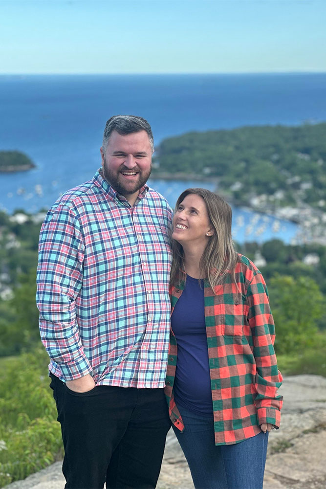 Andy and Emilee stand atop a hill overlooking a town and the ocean
