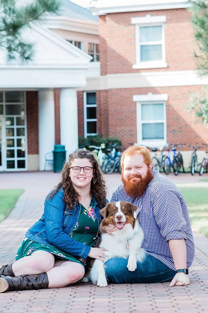 Victoria and Jacob sit on a brick pathway in front of a residence hall on CNU's campus with their dog.