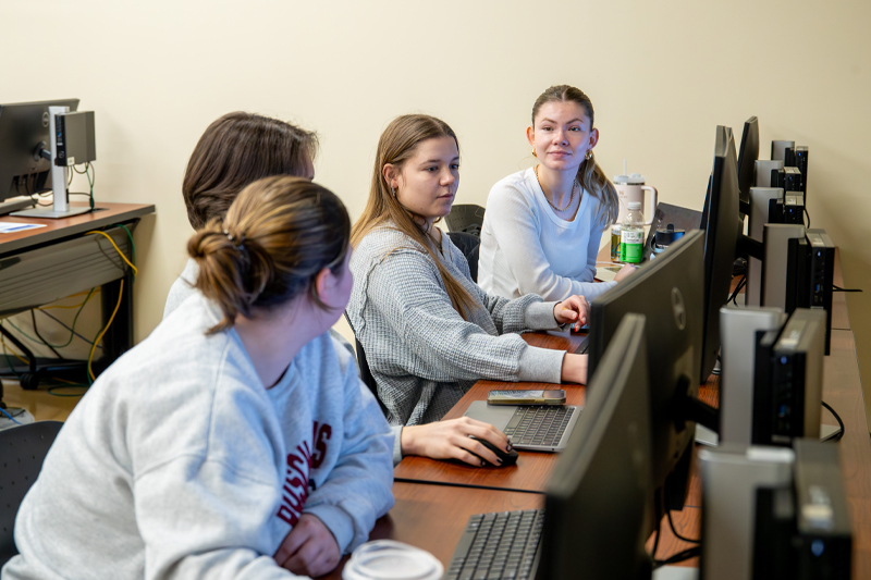 Students sit in front of computers in a lab while deep in discussion.