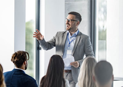 A man gestures and smiles while standing in front of a room full of business professionals.