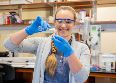 A student in a lab wearing safety googles and gloves pours a solution from a beaker into a container.