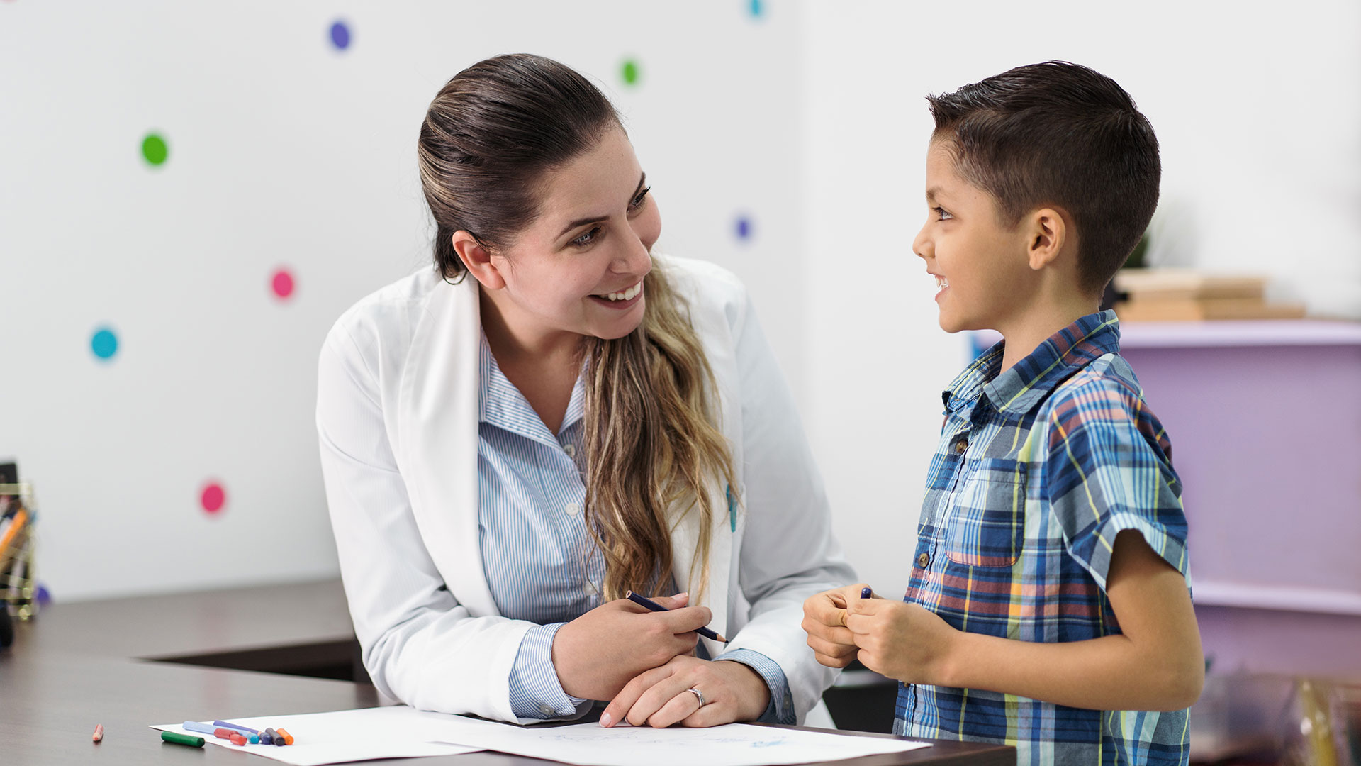 A woman works with a young child at a desk with paper on it.