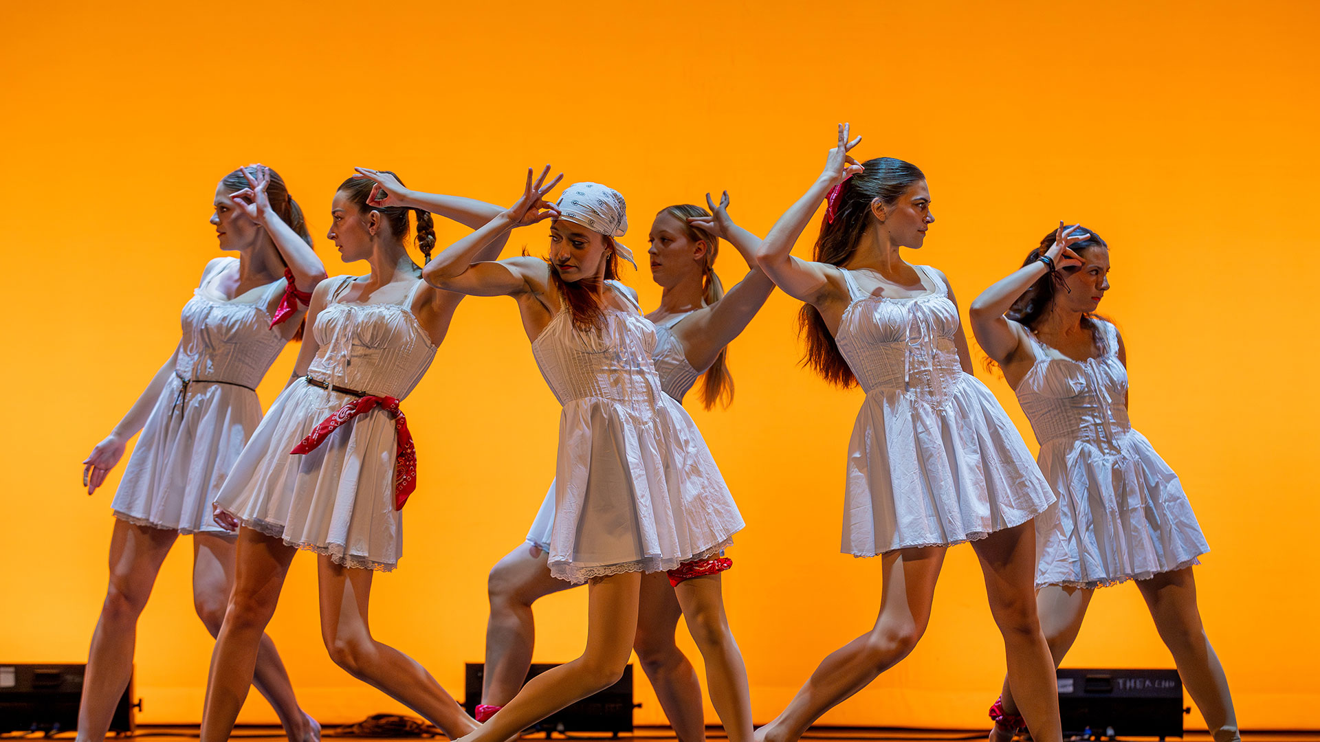 Six female dancers in white dresses and red accents, performing on stage with an orange background.