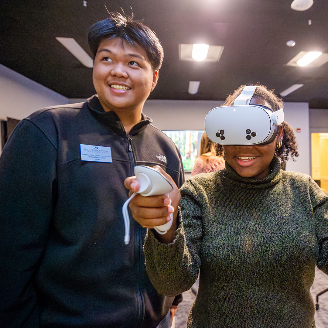 Two young adults are in a room with a black ceiling and bright lights. A Black woman is smiling and wearing a white virtual reality (VR) headset and holding two VR controllers, actively engaged in a VR experience. A man with a nametag is standing next to her, looking on and smiling.