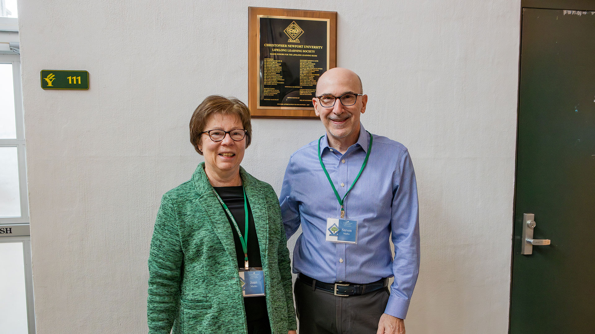Two members of the Lifelong Learning Society stand together in front of a plaque.