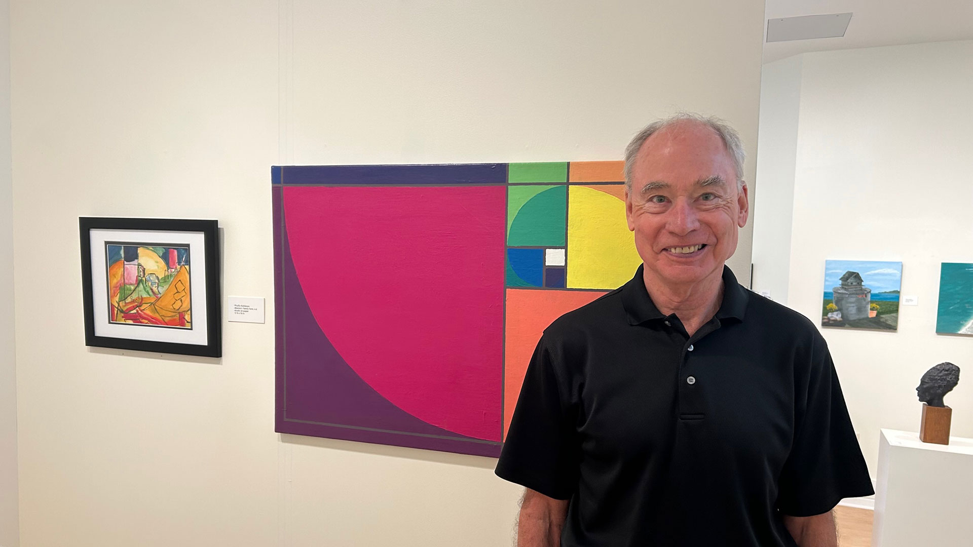 An older gentleman smiles in front of artwork hanging in a gallery at the Torggler Fine Arts Center