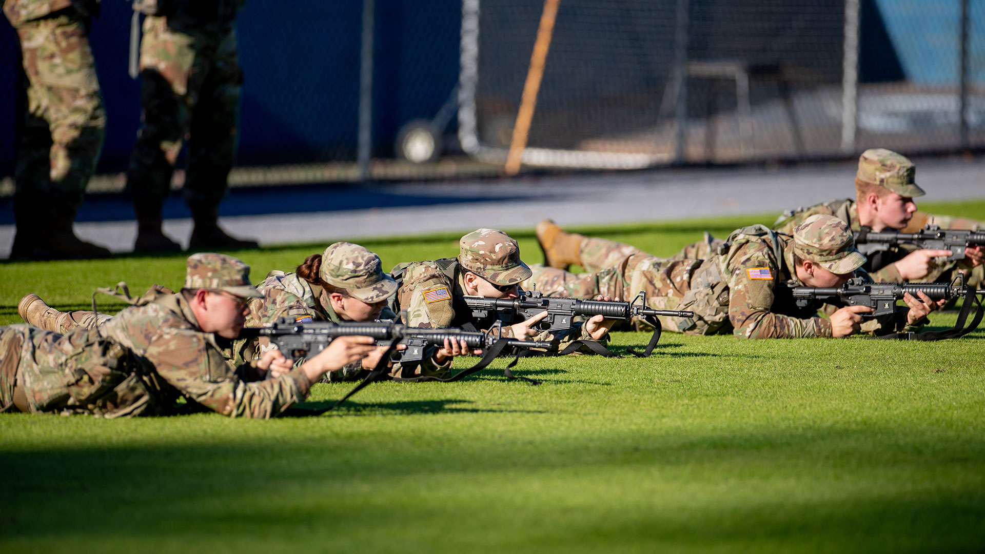 Four Army ROTC cadets in uniform practice marksmanship, lying prone on a grassy field with M4-style rifles.