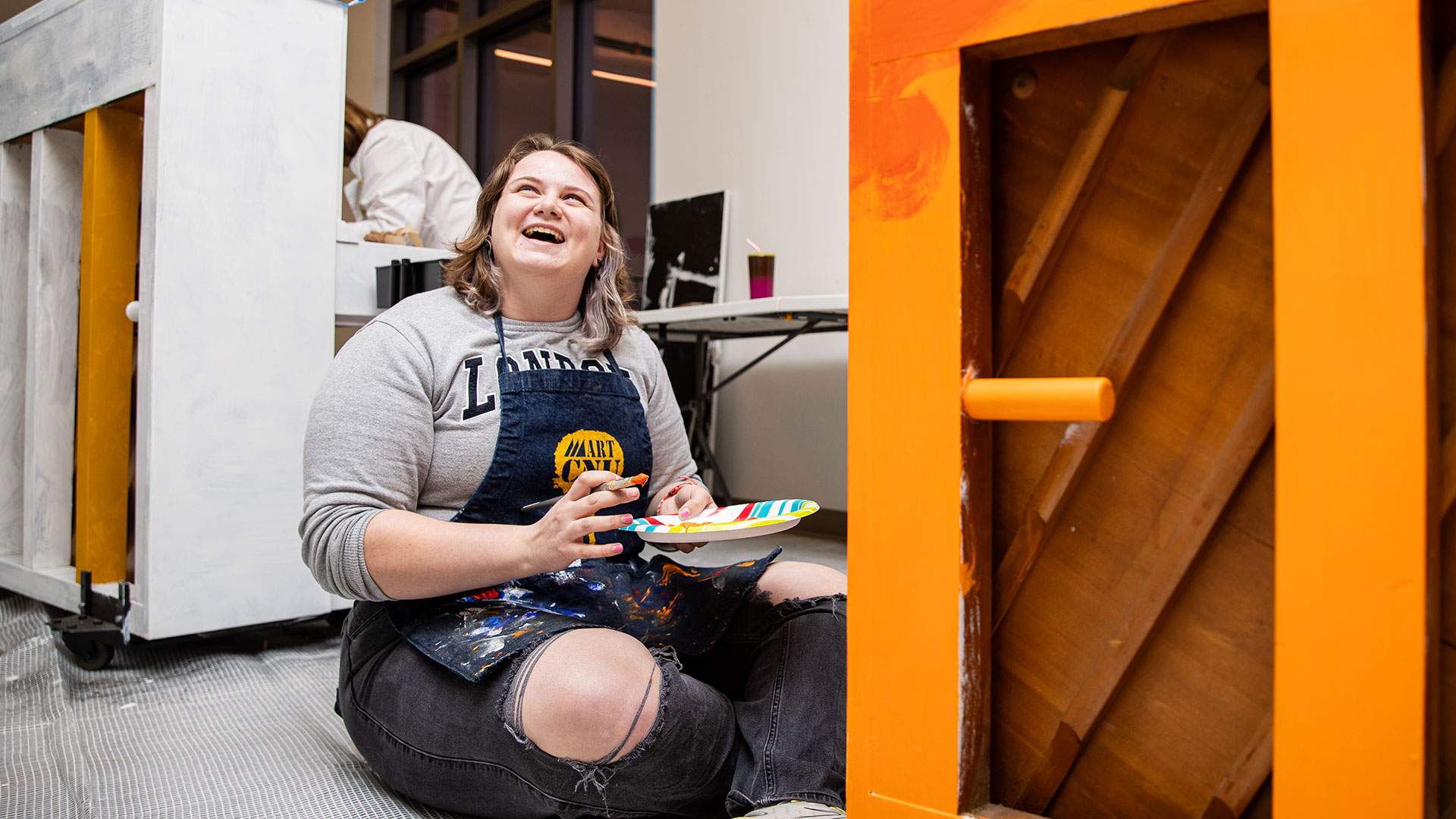 A young woman sits on the floor, smiling and looking up, as she paints a piano bright orange.