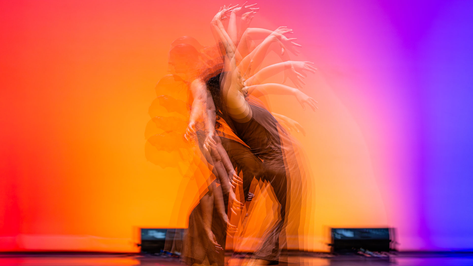 A dancer captured in a multiple-exposure photo, creating a blurred motion effect with their arms sweeping out, against a brightly lit stage backdrop that fades from orange and red on the left to purple and blue on the right. The text DANCE is in the lower right corner.