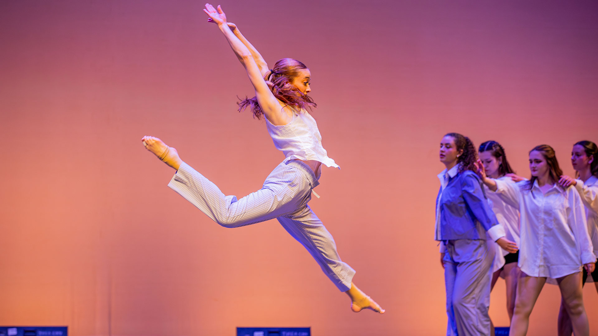 A female dancer is captured in a grand jete mid-air, wearing a white top and light blue-and-white striped pants. Her arms are stretched high above her head. Four other dancers are standing to the right on the stage, which is lit with purple and pink hues.