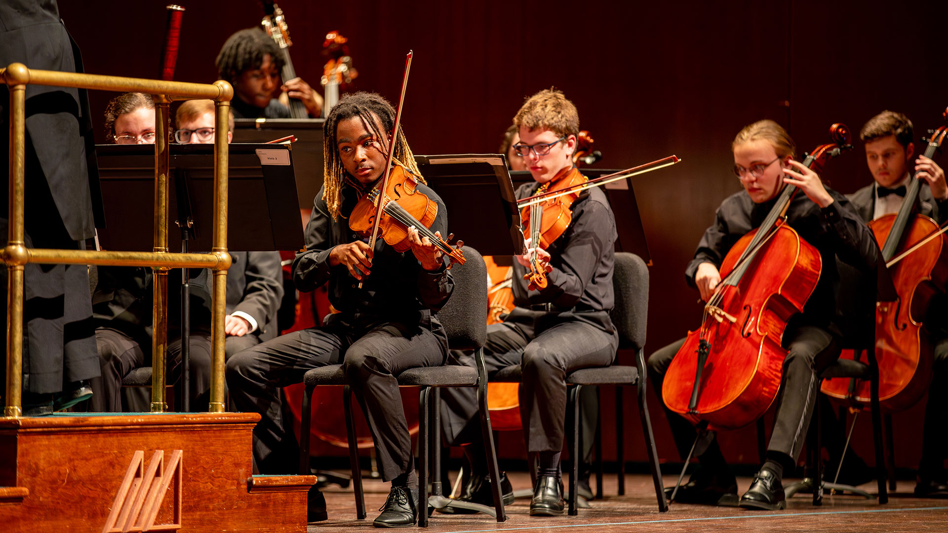A group of diverse young people in an orchestra, dressed in black, playing string instruments; the central focus is a Black male violinist with dreadlocks. The text MUSIC is in the lower right corner.